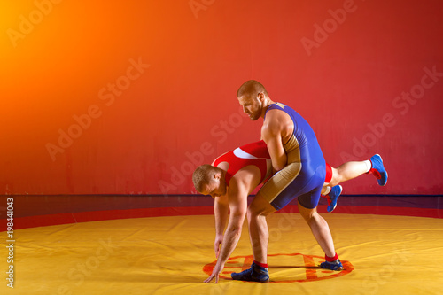 Two strong wrestlers in blue and red wrestling tights are wrestlng and making a making a hip throw on a yellow wrestling carpet in the gym. Young man doing grapple.