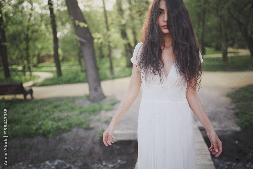 Lovely brooding brunette girl with long hair in white romantic dress in spring forest or park. The atmosphere of a warm sunny evening.