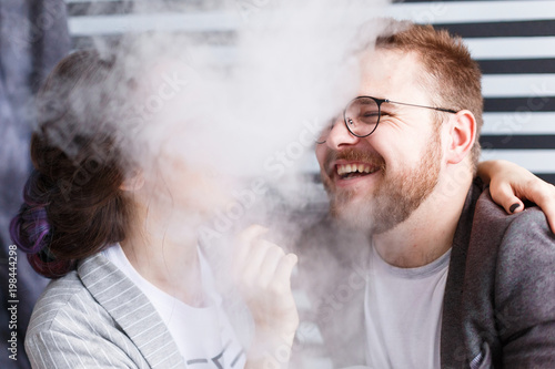 Couple hipsters vaping and smiling. Close-up portrait covered with smoke