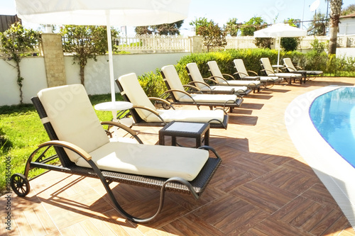 Beautiful tropical hotel resort with swimming pool, multiple sun-loungers deck chairs in row and palm trees on a warm sunny day. Clean water, spa treatments center. Close up, copy space, background.