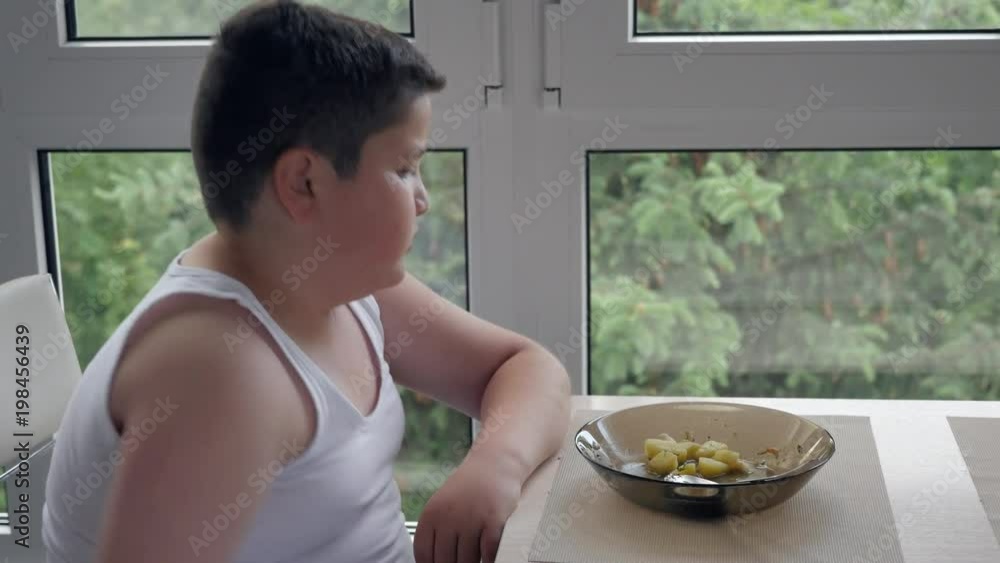 little fat boy sitting near the window and eats delicious soup. concept childhood obesity