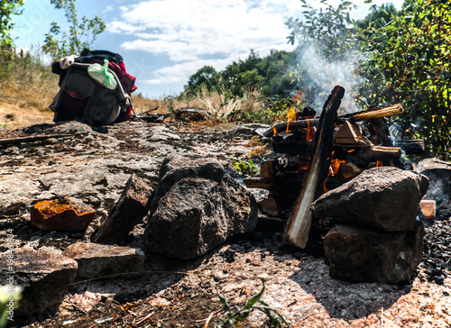 A view of a buning bonfire and a full touristic packpack. The photo was shot one summer day on the stone hill under a blue sky while hikers made a camp. photo