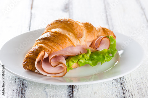 Fresh croissant with ham and salad leaf on white wooden background
