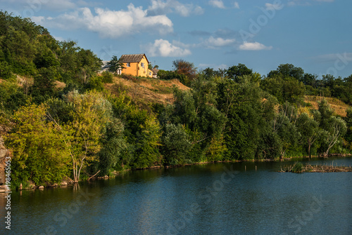 A beautiful summer view of a green hill with a small yellow house, located on the riverbank, Gordashovka village, Ukraine.