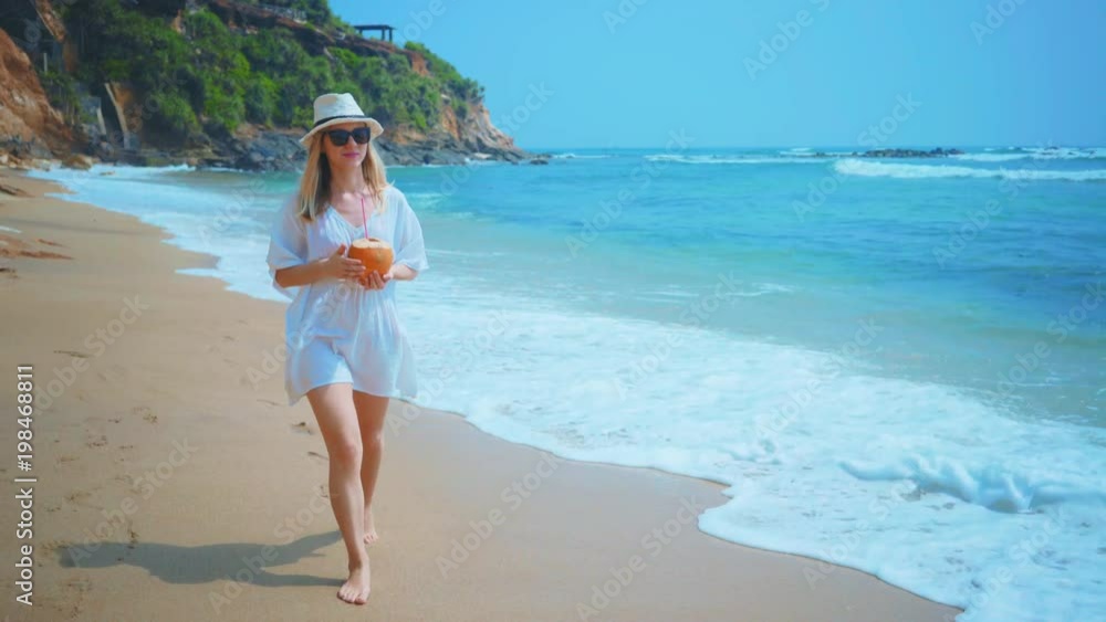Woman drinks coconut water and walking on the ocean beach under blue sky in sunny day. Seascape view with palms. Woman drinks coconut water and walking on the ocean beach under blue sky in sunny day. Seascape view with palms.