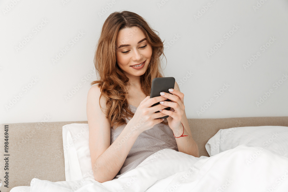Portrait of attractive woman resting in bed after sleep with white clean linen at bedroom and chatting online in cell phone Portrait of attractive woman resting in bed after sleep with white clean linen at bedroom and chatting online in cell phone