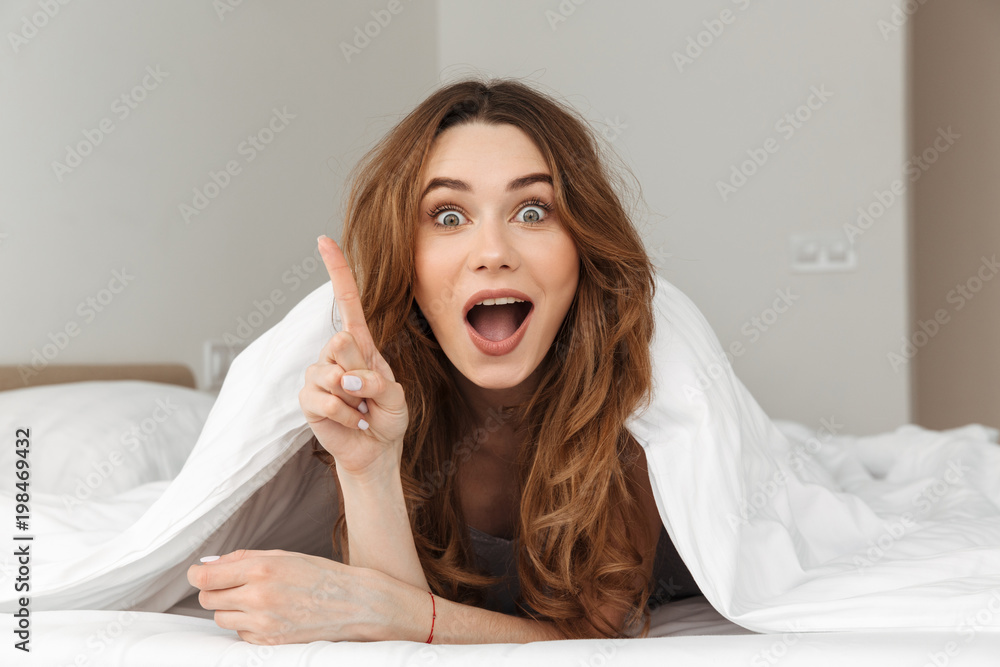 Photo of excited woman 20s lying in bed under white blanket in bedroom, and having idea putting index finger up Photo of excited woman 20s lying in bed under white blanket in bedroom, and having idea putting index finger up