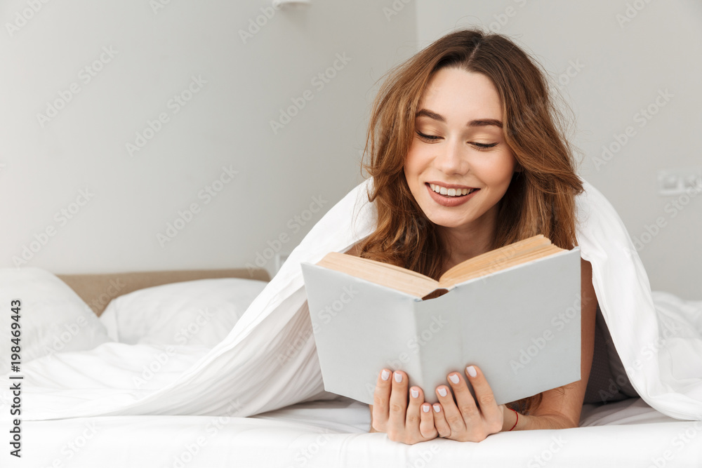 Portrait of joyous woman lying in bed under white blanket in bedroom, and reading interesting book Portrait of joyous woman lying in bed under white blanket in bedroom, and reading interesting book