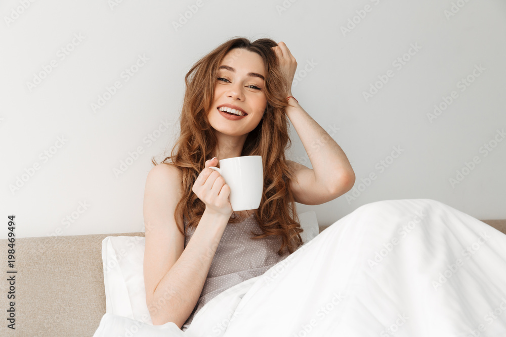 Portrait of fresh smiling woman resting after sleep in bed with white clean linen in bedroom, and drinking morning coffee or tea Portrait of fresh smiling woman resting after sleep in bed with white clean linen in bedroom, and drinking morning coffee or tea