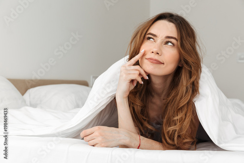 Portrait of smiling woman 20s lying in bed under white blanket in bedroom, and daydreaming or thinking looking upward