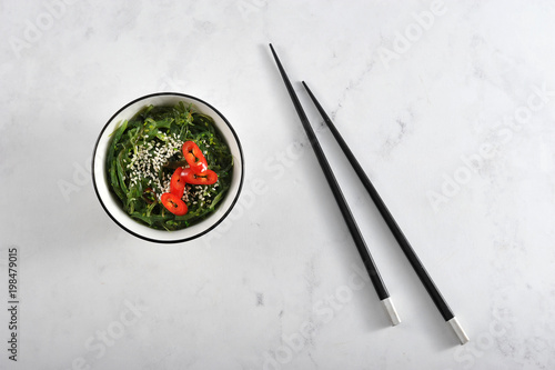 Salad of seaweed chuka in a round bowl. Salad is supplemented with sesame seeds and rings of hot chili peppers. In the frame, chopsticks. Light marble background. View from above. Close-up.