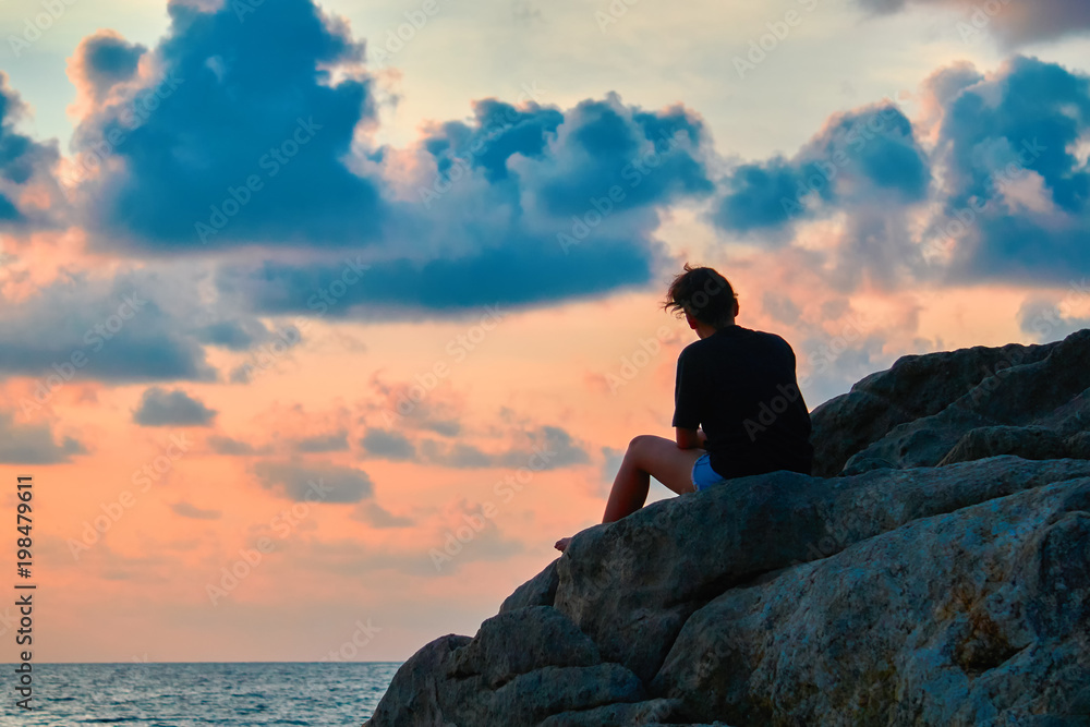 Woman silhouette at sunset. Girl sits on rock above sea. Evening sky of orange color with blue clouds. Seascape.