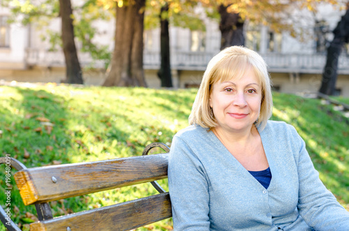   Middle-aged blond woman sitting on a  bench in the park looking at the camera on the autumn spring background. Portrait of beautiful senior happy elderly blond woman in a park on a sunny day.
