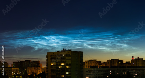 Wide panoramic night sky view of beautiful noctilucent clouds over the city with a cityscape roofs at foreground, rare phenomena which happens only on summer nights. cityline with awesome cloudy sky
