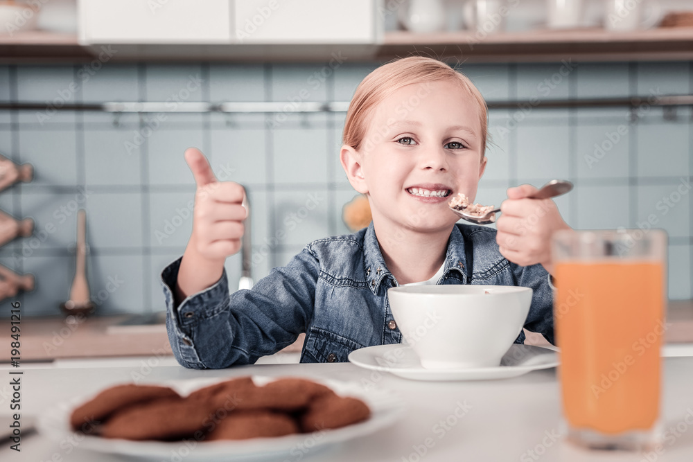 I recommend. Pleased child keeping smile on her face and raising thumb while looking at camera I recommend. Pleased child keeping smile on her face and raising thumb while looking at camera