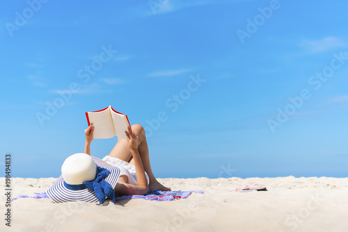 Fototapeta Naklejka Na Ścianę i Meble -  Young woman lying on the beach with reading a book on sand with blue sky. Happy and relaxing times on vacation.