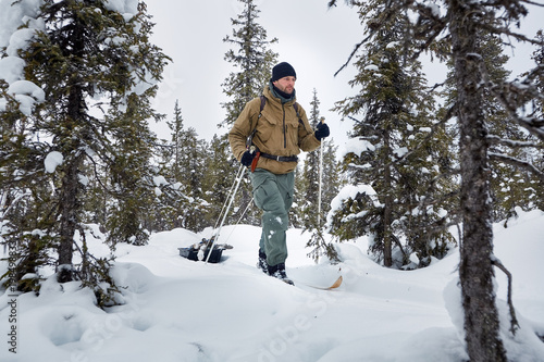 Through the forest. Young man in outdoors clothes is riding on wooden skis, on path in the forest and pulling sled across witer tundra