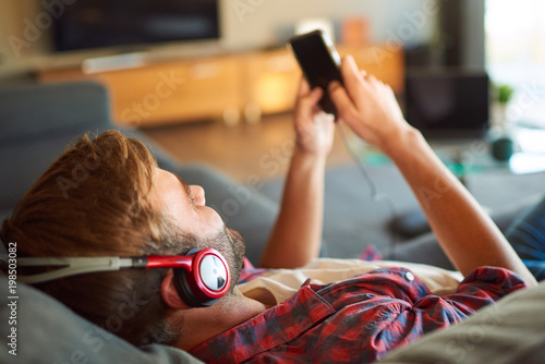 Over the shoulder image of a young caucasian guy listening to music through headphones from his phone that he is holding in his hands, all while relaxing in his lovley modern living room. photo