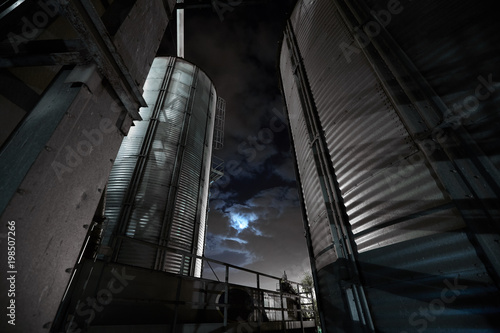 Modern grain terminal at night. Metal tanks of elevator. Grain-drying complex construction. Commercial grain or seed silos at seaport. Steel storage for agricultural harvest.