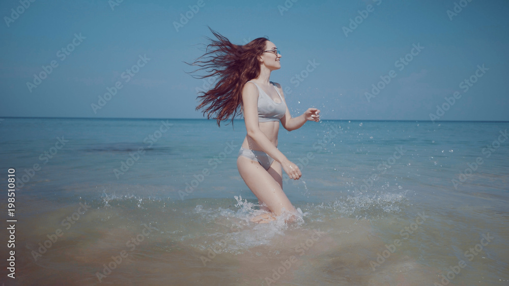 Naklejka premium Beautiful happy young girl in bikini and sunglasses playing in the sea water over sky background during summer holiday