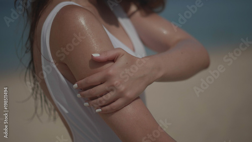 Beautiful girl in swimsuit applying sunscreen lotion tan oil on body on the tropical sandy beach during summer holiday over sea and sky background