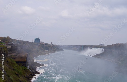 Beautiful and impressive panorama of the Niagara Falls in Ontario (Canada) on a bright autumn day with water crashing down the falls onto rocks creating lots of mist in front of the city skyline