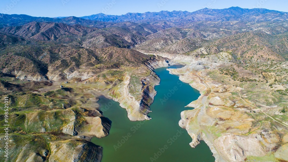 Fototapeta premium Aerial bird eye view of Kalavasos rockfill dam, Larnaca, Cyprus. The Vasilikos river streaming towards the reservoir and the hills around the water from above.