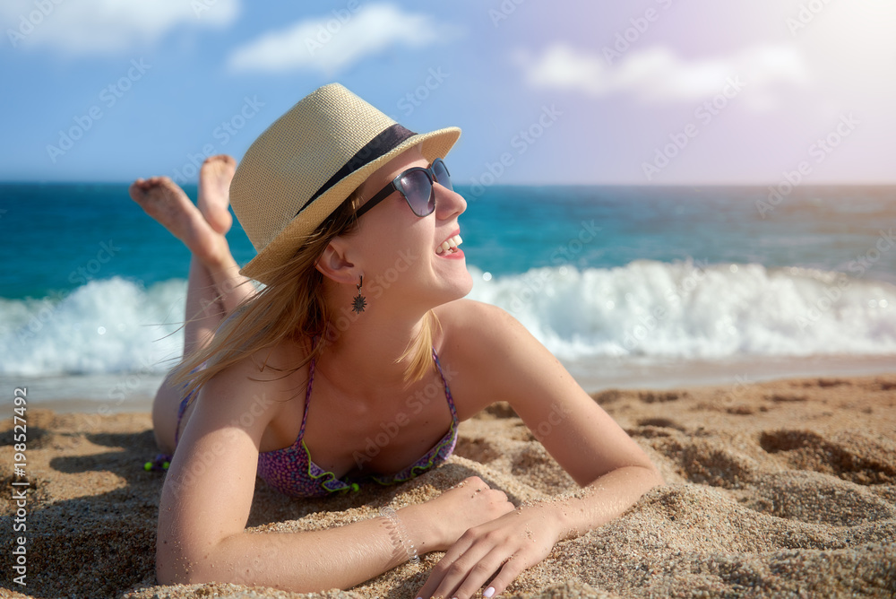 Portrait of a young woman in sunhat and swimsuit is laying on the beach sand against the sea.