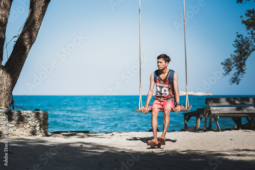 Young man sits on a rope swing on sandy shore against blue sea and clear sky. Asian man wears T-shirt with 