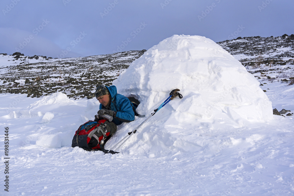 hiker pours himself a tea from a thermos, sitting in a snowy hut igloo against a background of a winter mountain landscape..