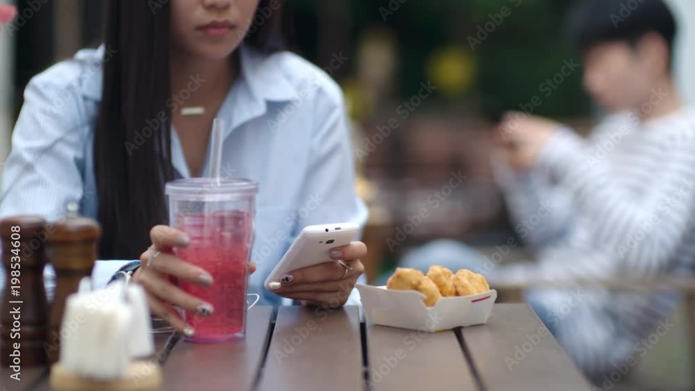 Tracking shot of young Asian woman sitting at cafe table outdoors, drinking soda and texting on smartphone at lunch Tracking shot of young Asian woman sitting at cafe table outdoors, drinking soda and texting on smartphone at lunch
