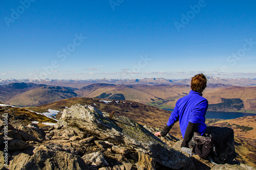 A male hiker sits upright and rests and looks in to the distance on a clear bright sunny day at the view of the Scottish Highlands from summit of Ben More, Trossachs