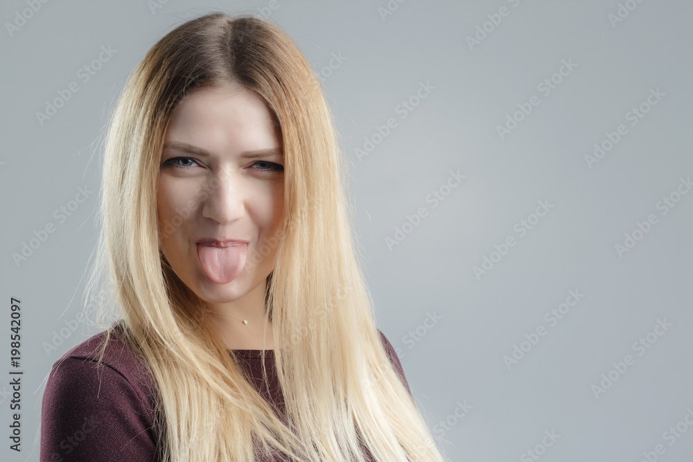 Fototapeta premium Beautiful girl thumbs up, tongue out and looking at camera. Indoor shot, gray background