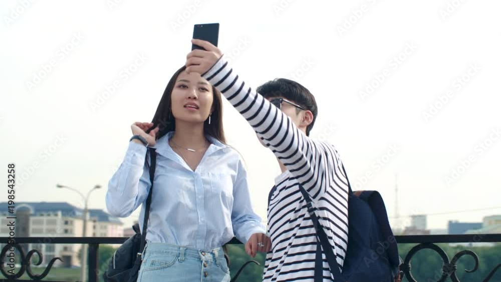 Teenage Asian boy and girl in trendy outfits standing at riverside in the city at summer day and taking selfie with smartphone Teenage Asian boy and girl in trendy outfits standing at riverside in the city at summer day and taking selfie with smartphone