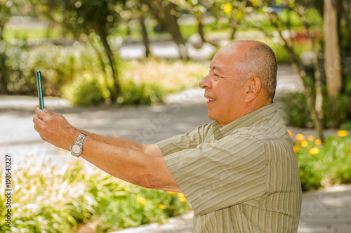 Outdoor view of old man sits on bench doing video cam with cellphone surrounding of nature. All problems left behind