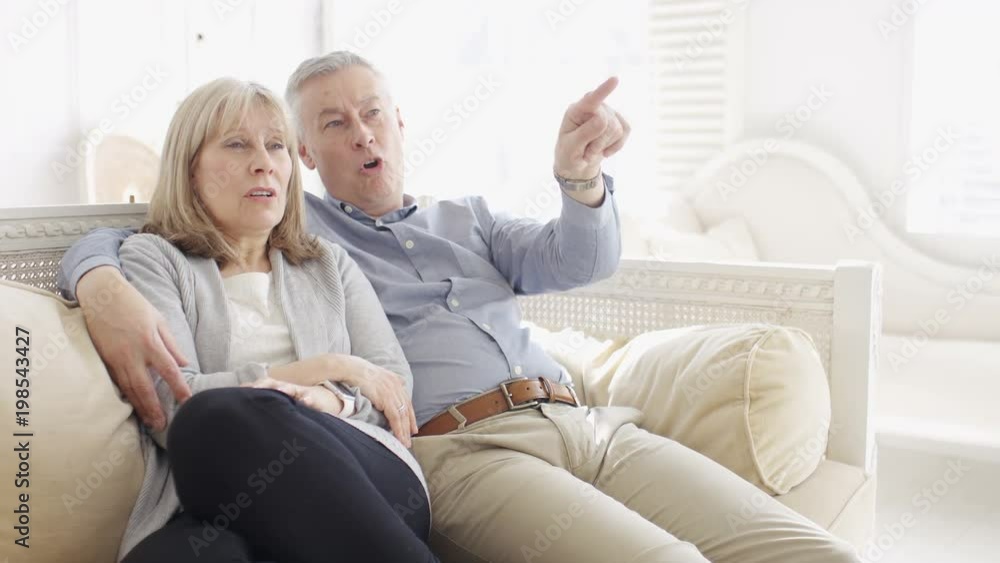 Mature couple watching tv as the husband points out something he doesn't like Mature couple watching tv as the husband points out something he doesn't like