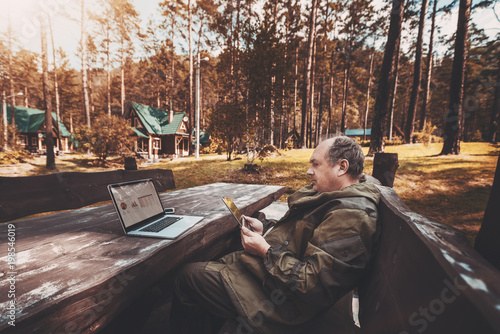 Aged businessman in a dark-green overall is sitting on the wooden bench at the table being on leave and using the digital tablet for video call with his colleagues; laptop with a graph on the table