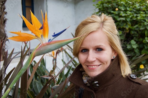 Pretty blonde female European tourist in San Diego, California (USA) next to a large Strelitzia / bird of paradise flower in the historical city center on a bright winter day	