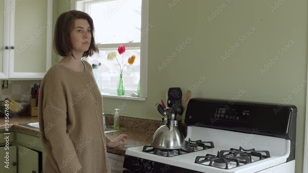 Woman standing by stove looking into the distance as she waits for her teapot to heat up. Woman standing by stove looking into the distance as she waits for her teapot to heat up.