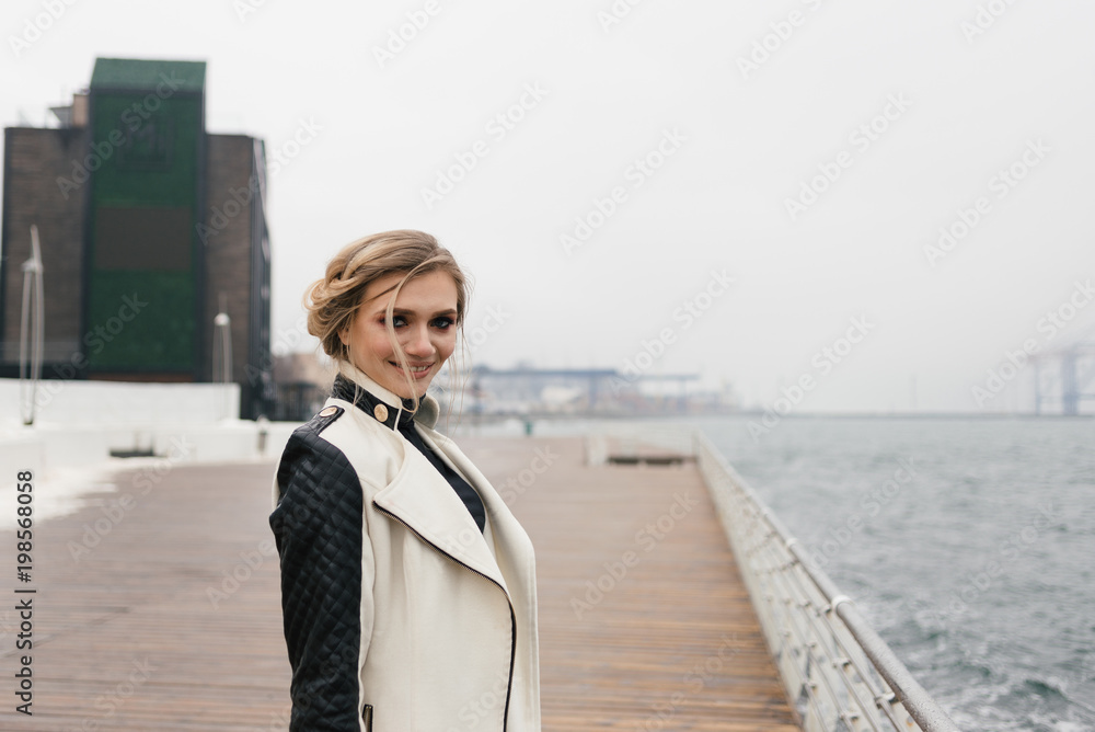 Beautiful cheerful student standing on the waterfront keeps herself by the head.