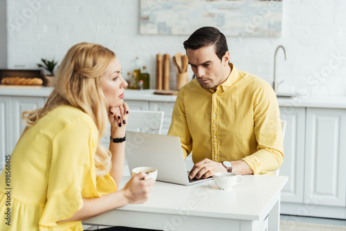 Принти на полотні Sad girlfriend holding coffee and sitting by her boyfriend working on laptop in