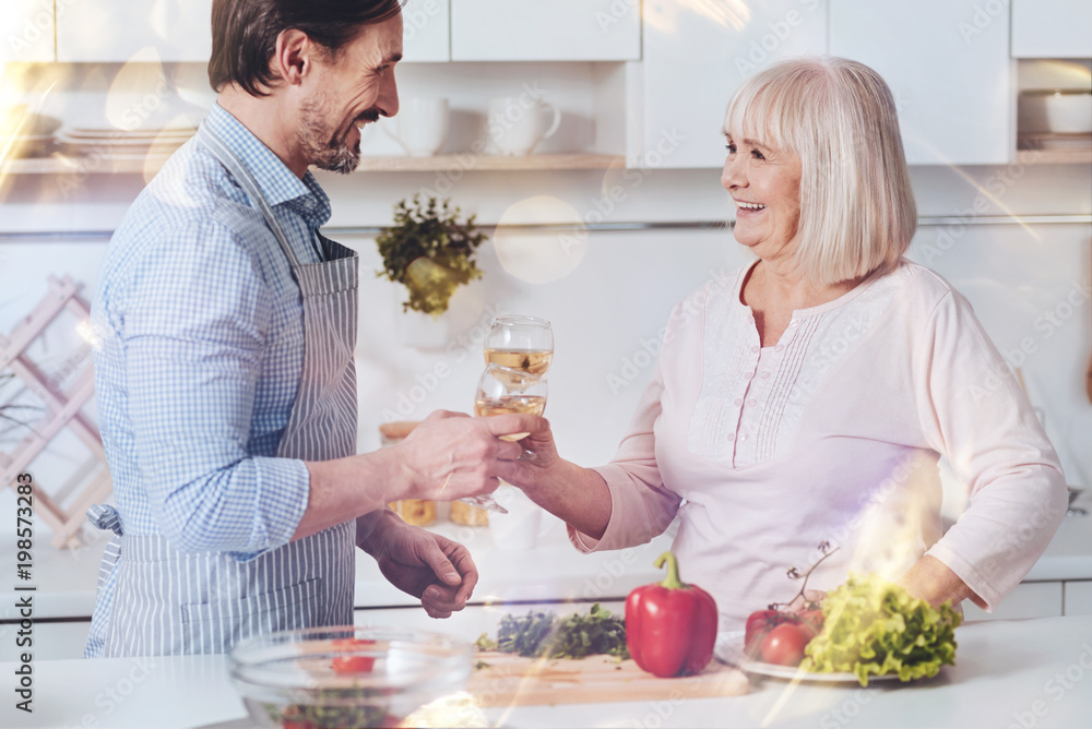 For you. Cheerful delighted aged woman and her adult son drinking wine and standing in the kitchen while enjoying time together