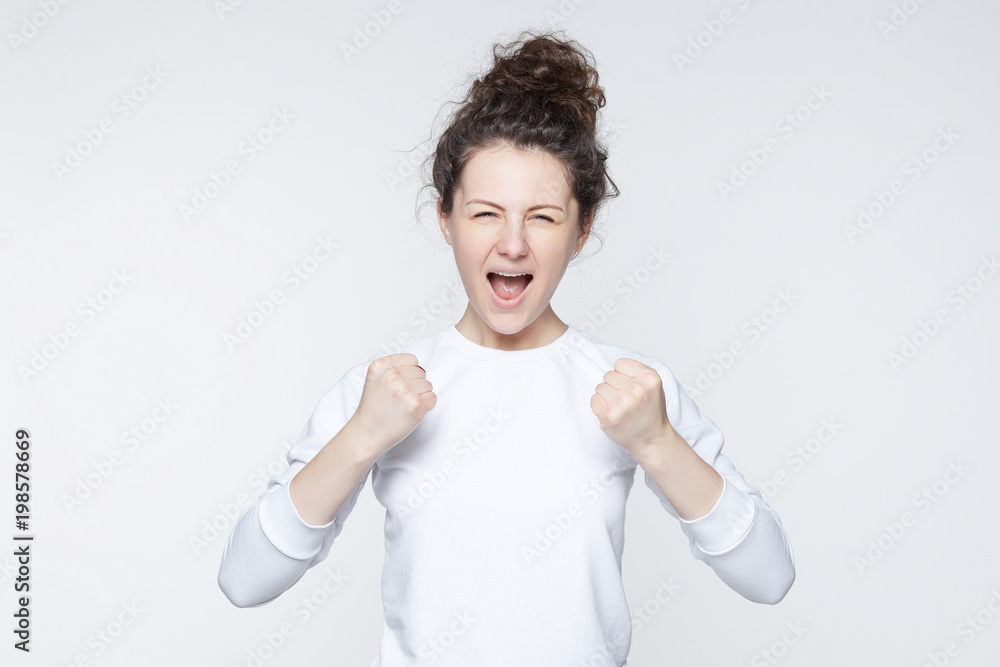 Waist up portrait of strong successful determined young female winner in casual t-shirt raising arms, clenching fists, exclaiming with joy and excitement. Victory, success and achievement concept. Waist up portrait of strong successful determined young female winner in casual t-shirt raising arms, clenching fists, exclaiming with joy and excitement. Victory, success and achievement concept.