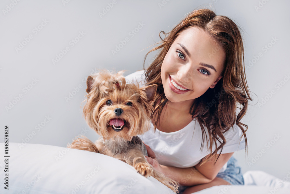 portrait of young smiling woman with yorkshire terrier resting on bed isolated on grey portrait of young smiling woman with yorkshire terrier resting on bed isolated on grey
