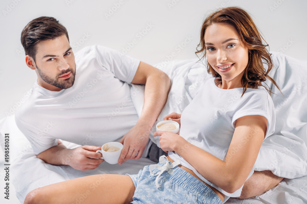 Fototapeta premium young couple with cups of coffee resting in bed together isolated on grey