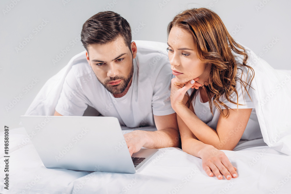 portrait of focused couple using laptop while resting on bed together isolated on grey portrait of focused couple using laptop while resting on bed together isolated on grey