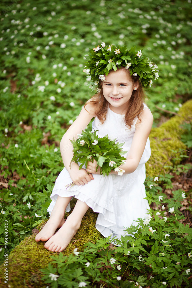 Beautiful little girl in a white dress walks in the spring wood. Portrait of the pretty little girl with a wreath from spring flowers on the head. Beautiful little girl in a white dress walks in the spring wood. Portrait of the pretty little girl with a wreath from spring flowers on the head.