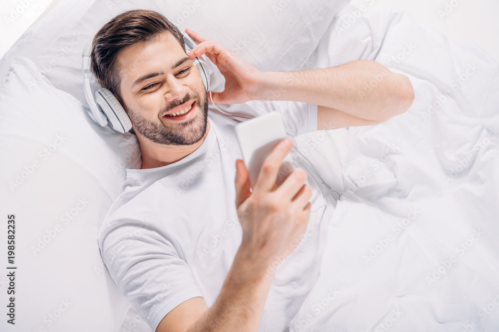 Obraz premium overhead view of smiling man in headphones using smartphone in bed