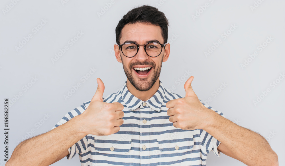 Portrait of handsome male with healthy toothy smile, wears round trendy glasses, shirt looking at the camera with happy expression, showing thumbs up with both hands. Body language and emotion concept Portrait of handsome male with healthy toothy smile, wears round trendy glasses, shirt looking at the camera with happy expression, showing thumbs up with both hands. Body language and emotion concept