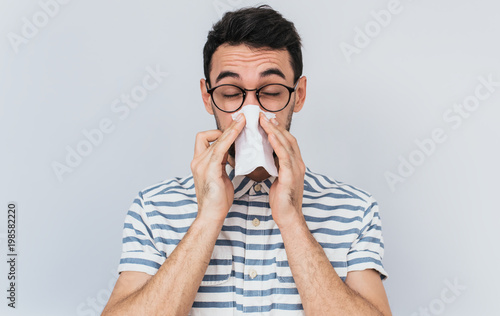Fototapeta Naklejka Na Ścianę i Meble -  Horizontal portrait of unhealthy handsome man wearing striped shirt and glasses, blowing nose into tissue. Male have flu, virus or allergy against white background. Healthy medicine and people concept